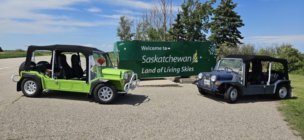 Two Mini Mokes sitting in front of a "Welcome to Saskatchewan" sign on the Saskatchewan and Manitoba border.