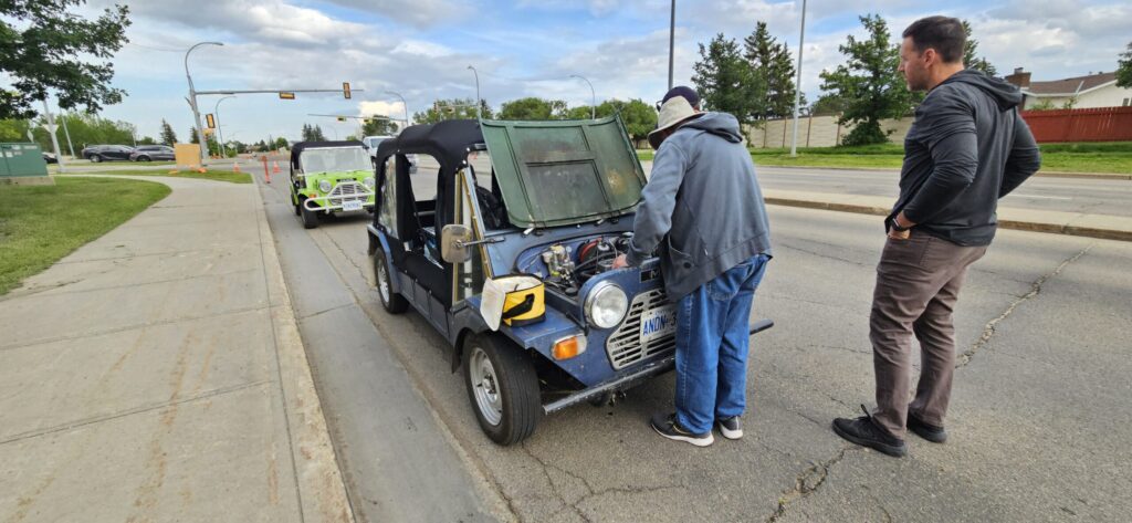 A Blue Mini Moke is at the side of the road with it's engine hood open and guys standing around trying to figure out what is wrong